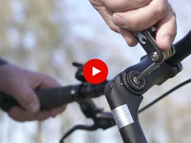 Nahaufnahme von Händen, die den Fahrradlenkervorbau mit einem Inbusschlüssel einstellen. Roter Play-Button zeigt Video-Tutorial-Inhalt an.