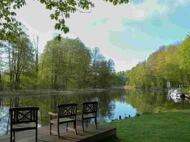 Drei Holzstühle auf einem Steg mit Blick auf einen ruhigen See, umgeben von grünen Bäumen. Ein Segelboot liegt am rechten Ufer unter blauem Himmel.