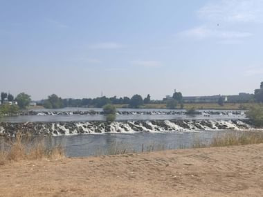 Weite Ansicht des Ruhr-Flusses mit einem Stufenwehr, das weiße Wasserkaskaden erzeugt. Sandiges Flussufer im Vordergrund, Industriegebäude in der Ferne.