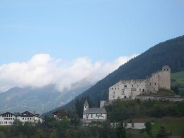 Mittelalterliche Burg Heinfels auf einem Hügel mit weißer Kirche und Dorfhäusern darunter, umgeben von bewaldeten Alpenbergen unter bewölktem Himmel.