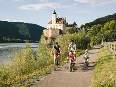 Vierköpfige Familie radelt auf Weg neben der Donau mit historischer Burg auf Hügel. Grüne Hügel und bewölkter Himmel im Hintergrund.