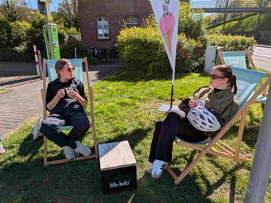 Two women relaxing in deck chairs on grass, enjoying ice cream. Cycling helmets rest nearby. Pink ice cream flag and brick building in background.