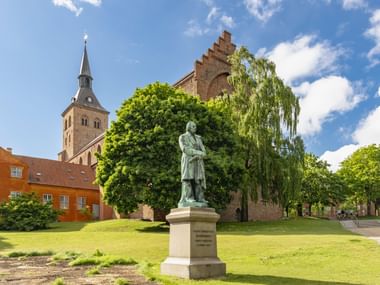 Bronze statue of Hans Christian Andersen on stone pedestal in park with historic brick church tower and buildings in background under blue sky.