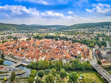 Luftaufnahme von Hann. Münden mit historischer Altstadt mit roten Ziegeldächern, Fluss, Brücke und bewaldeten Hügeln unter blauem Himmel.