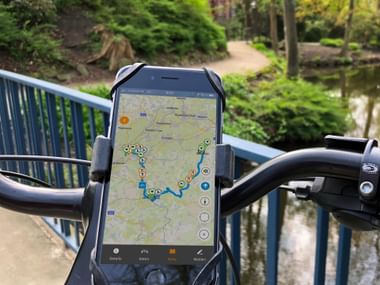 iPhone mounted on bicycle handlebars displaying cycle route navigation app with blue route line and location markers on map.