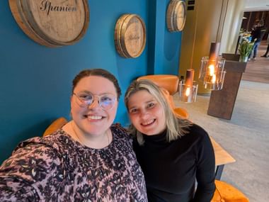 Zwei lächelnde Frauen sitzen an einem Tisch in einem modernen Interieur mit blauen Wänden, die mit Holzweinfässern dekoriert sind, und orangen Stühlen.