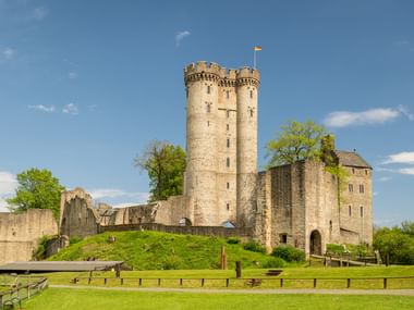 Medieval Kasselburg castle ruins with twin towers and stone walls on a grassy hill, surrounded by wooden fencing under blue sky.