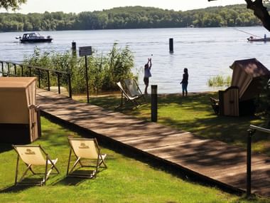 Lakefront terrace with wooden deck, beach chairs, and wicker beach basket overlooking a lake with boats and forested hills in Potsdam.
