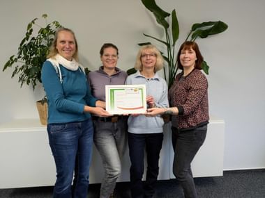 Four smiling women standing together holding a framed certificate. Green plants are visible in the background against a white wall.