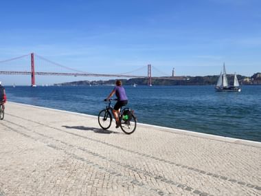 Radfahrer auf gepflasterter Uferpromenade mit roter Hängebrücke über blauem Wasser, Segelboot in der Ferne unter klarem Himmel.