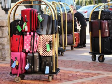 Mehrere goldene Gepäckwagen beladen mit bunten Koffern in Rot, Pink, Schwarz und Punktmustern mit Gepäckanhängern auf Ziegelpflaster.