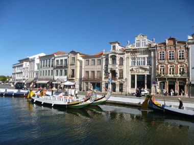 Blick vom Wasser auf Aveiros historische Uferpromenade mit bunten traditionellen portugiesischen Booten vor prächtigen Gebäuden bei blauem Himmel.