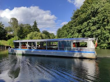 Blue and white passenger boat named Solaaris with solar panels on roof cruising on calm river surrounded by lush green trees under blue sky.