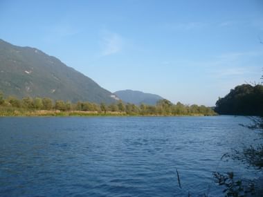 Idyllischer Blick auf die Drau mit ruhigem blauen Wasser, grüner Vegetation entlang der Ufer und bewaldeten Bergen unter blauem Himmel.