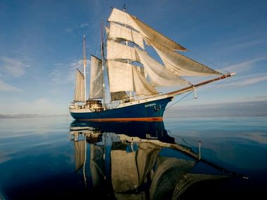 Blau-rumpfiges Segelschiff Antigua mit weißen Segeln, spiegelt sich in ruhigem Wasser unter klarem Himmel mit Bergen am Horizont.