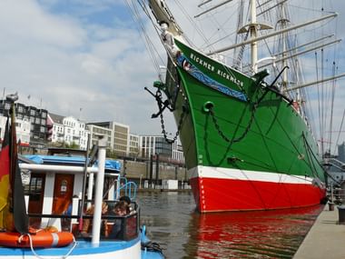 Grünes und rotes historisches Segelschiff mit hohen Masten im Hamburger Hafen, mit modernen Gebäuden und deutscher Flagge im Hintergrund.