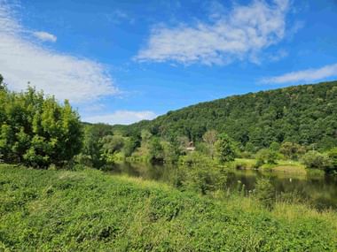 Ruhiger Fluss schlängelt sich durch üppige grüne Landschaft mit dicht bewaldeten Hügeln unter blauem Himmel mit vereinzelten Wolken.