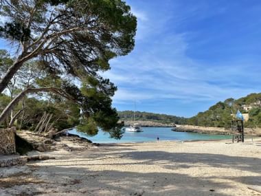 Sandstrand im Naturpark Mondragon mit türkisfarbenem Wasser, Pinien, verankertem Segelboot und Rettungsturm unter blauem Himmel.