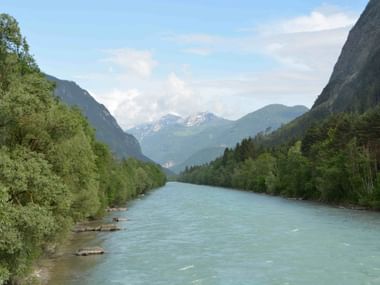 Türkisfarbene Drau fließt durch ein Tal zwischen Lienz und Oberdrauburg, mit bewaldeten Hängen und schneebedeckten Bergen in der Ferne.