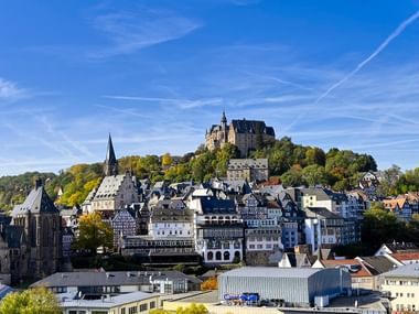 Panoramablick auf Marburg Oberstadt mit historischem Schloss auf dem Hügel, gotischem Kirchturm und traditionellen Fachwerkhäusern unter blauem Himmel.