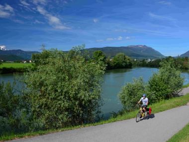 Radfahrer mit Packtaschen auf asphaltiertem Weg neben der Drau bei Villach. Grüne Ufer, Bäume und Berge unter blauem Himmel sichtbar.