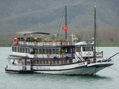 Vietnamesisches Touristenboot in Garden Bay Weißes mehrstöckiges Touristenboot mit vietnamesischer Flagge fährt in den Gewässern der Garden Bay, Berge im Hintergrund sichtbar.