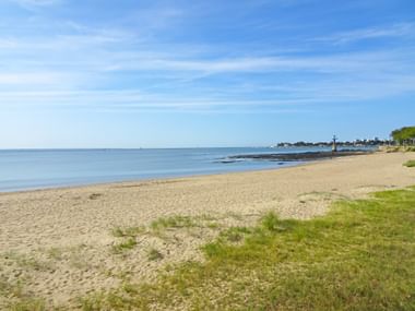 Breiter Sandstrand in St-Nazaire mit ruhigem blauen Meer, grünem Gras im Vordergrund und Küstengebäuden in der Ferne unter blauem Himmel.
