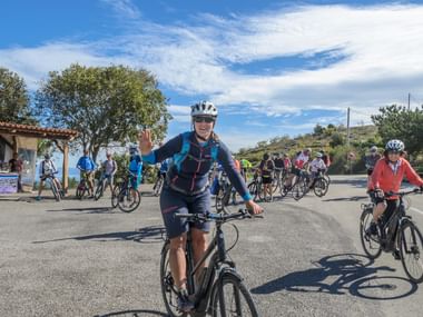 Große geführte Radgruppe auf asphaltierter Straße unter blauem Himmel. Führender Radfahrer in blauer Jacke winkt beim Fahren, viele Teilnehmer folgen.