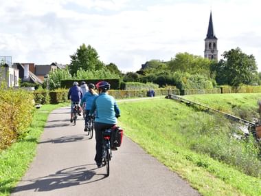 Cyclists on path near Sint-Amands church Group of cyclists with panniers riding on paved path through green countryside near Sint-Amands, with church tower visible in background.