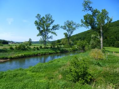 Werra-Flusslandschaft bei Bad Salzungen Malerischer Blick auf die Werra, die durch grüne Landschaft mit üppigen Wiesen, Bäumen und bewaldeten Hügeln unter blauem Himmel fließt.