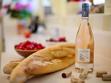 Bottle of Ventoux rosé wine from La Verrière beside fresh baguettes, wine corks, and a bowl of tomatoes on a bright kitchen counter.