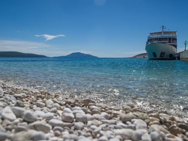 White ferry moored at pier in Kvarner Bay, Croatia. Crystal clear turquoise water laps against pebble beach with mountains in background.