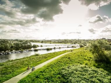 Aerial view of Hattingen Ruhr Valley showing cyclists on a path beside the Ruhr River, surrounded by green meadows and trees under a cloudy sky.