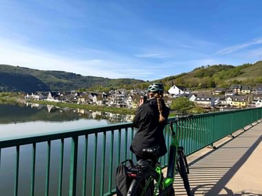 Radfahrer auf grüner Brücke über die Mosel bei Karden, mit Flussdorf und Hügeln, die sich im ruhigen Wasser unter blauem Himmel spiegeln.