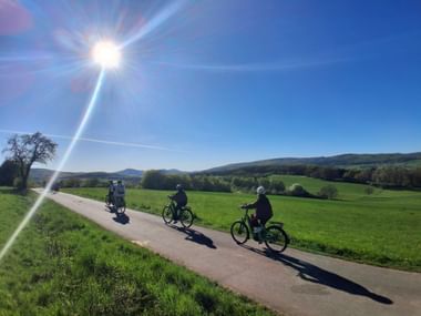 Three cyclists riding on a paved path through green meadows in the Marburg countryside under bright sunshine with rolling hills in the background.