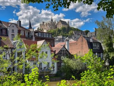 View of Marburg's historic town center with traditional half-timbered houses along the river, dominated by the medieval castle on the hill above.