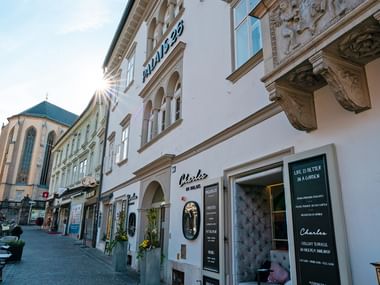 Historisches weißes Gebäude mit ornamentaler Barockfassade und Hotel Palais 26 Beschilderung. Erdgeschossläden säumen die Kopfsteinpflasterstraße in Villach.