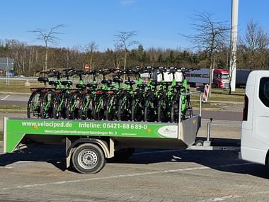 Green Velociped trailer loaded with multiple bicycles attached to a white van, parked on a street with trees and buildings in the background.