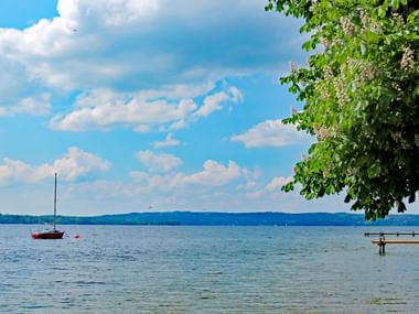 View of Lake Starnberg with sailboat Scenic view of Lake Starnberg with a red sailboat anchored on calm blue water. Green tree branches frame the top right, blue sky with white clouds above.