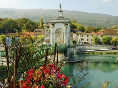 Green suspension bridge crossing the turquoise Rhône River in Seyssel with red flowers in foreground and mountains in background.