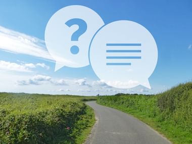Zwei Sprechblasen mit Fragezeichen und Textzeilen über einer schmalen asphaltierten Straße durch grüne Landschaft unter blauem Himmel mit Wolken.