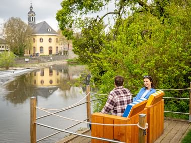 Couple sitting in wooden chairs on riverside deck overlooking Lahn River in Wetzlar, with historic church and buildings reflected in water.