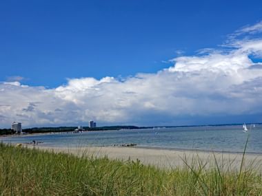 Sandiger Ostsee-Strand mit Grasdünen im Vordergrund, ruhiges blaues Wasser mit weißen Segelbooten und Küstengebäuden unter bewölktem Himmel.