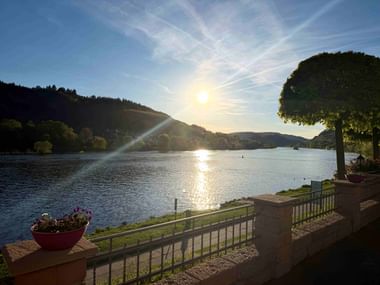 Blick von Hotelterrasse mit Blumentöpfen und Geländer auf die Mosel bei Sonnenuntergang, mit Hügeln und Bäumen auf beiden Seiten.