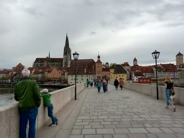Steinerne Brücke in Regensburg mit Fußgängern Richtung Altstadt. Domturm und historische Gebäude unter bewölktem Himmel sichtbar.
