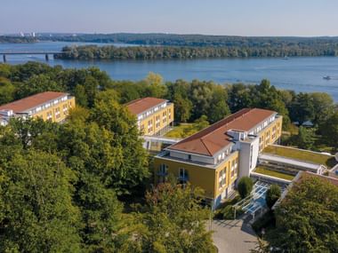 Luftaufnahme des Seminaris Seehotel Potsdam mit gelben Gebäuden umgeben von Bäumen mit Blick auf einen großen See mit Brücke im Hintergrund.