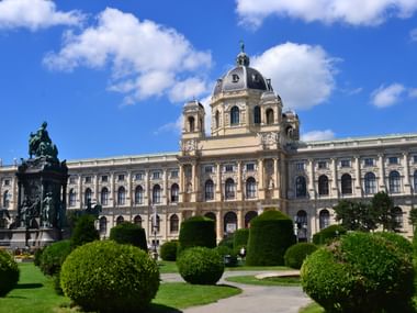 Naturhistorisches Museum Wien mit prachtvoller Barockfassade, zentraler Kuppel und Denkmal. Gepflegte Gärten mit runden Buchsbäumen im Vordergrund.