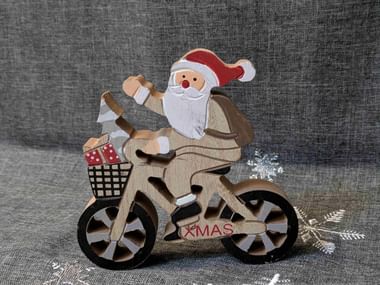 Wooden Christmas decoration showing Santa Claus riding a bicycle with gifts in the basket. The word XMAS appears on the frame against gray fabric.