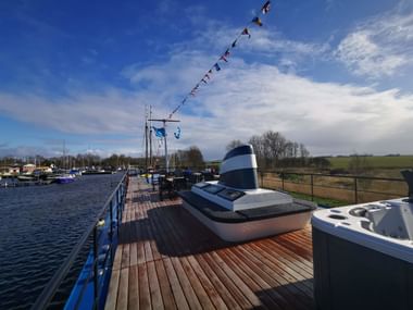 Sonnendeck der MS Magnifique mit Holzplanken, weißem Schornstein und blauen Geländern mit Blick auf Marina mit Booten unter bewölktem Himmel.