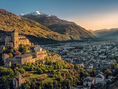Historic castle on a hill overlooking Sion in Valais at sunset, with snow-capped mountains and the Rhone valley in the background.